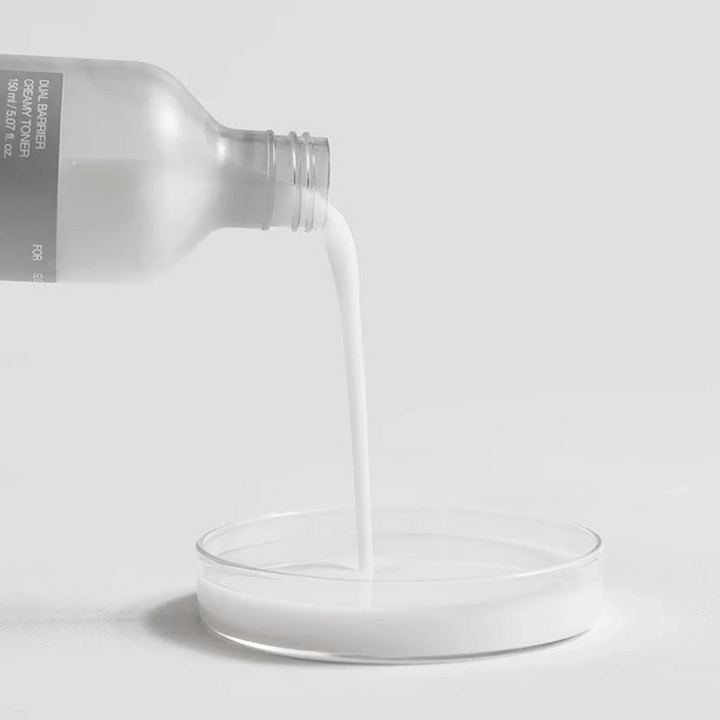 Celimax Dual Barrier Creamy Toner se previdno vlije iz steklenice v okroglo laboratorijsko posodo, pri čemer je njegova mlečna in hranljiva formula jasno v ospredju.