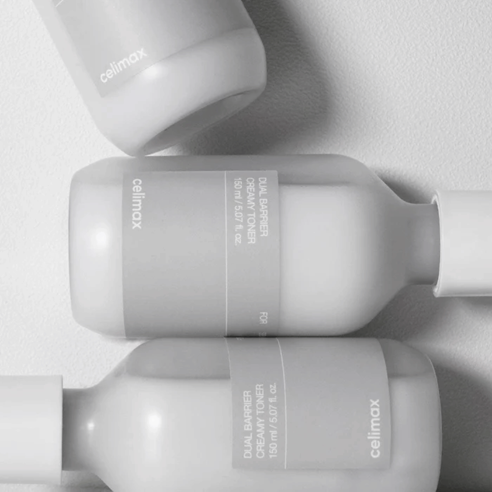 Tri ležeče steklenice Celimax Dual Barrier Creamy Toner v minimalističnem dizajnu s sivo etiketo in mehko svetlobo – popolno za občutljivo in suho kožo.