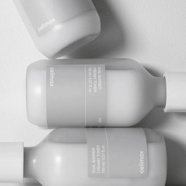 Tri ležeče steklenice Celimax Dual Barrier Creamy Toner v minimalističnem dizajnu s sivo etiketo in mehko svetlobo – popolno za občutljivo in suho kožo.