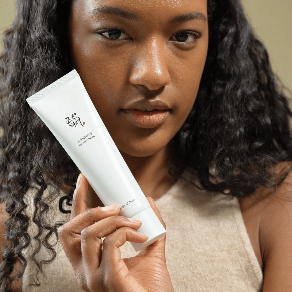 Ženska s kodrastimi lasmi drži tubo "Beauty of Joseon Dynasty Cream" blizu svojega obraza, kar poudarja izdelek. Krema, predstavljena v elegantni beli tubi, je zasnovana za hranjenje in pomlajevanje kože, kar poudarja njeno privlačnost za različne tipe kože.