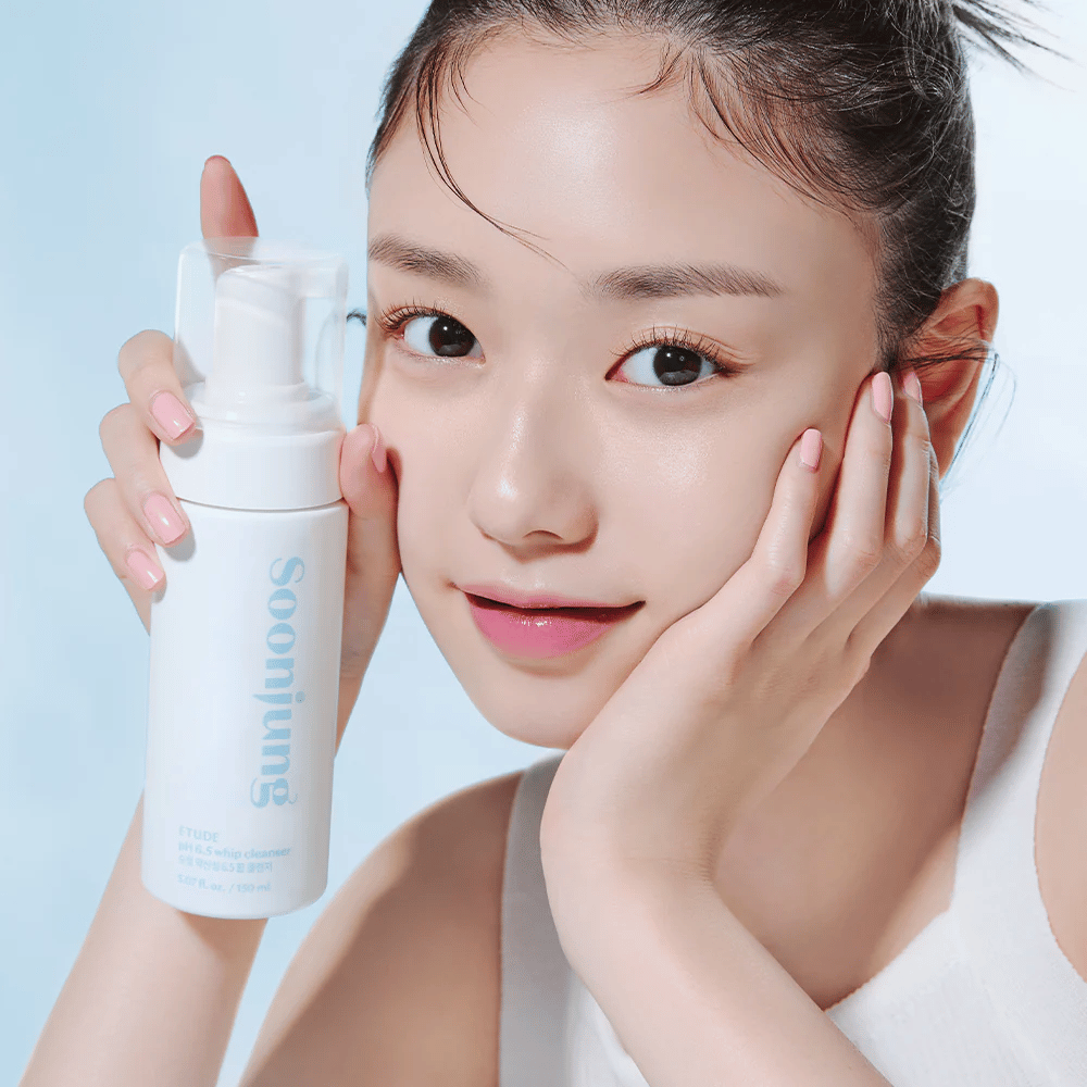 Mlada ženska s čisto kožo drži belo stekleničko s črpalko z napisom "Soon Jung pH 6.5 Whip Cleanser" od ETUDE. Nasmejana je proti kameri z lahkim ličenjem in svežim videzom na svetlo modrem ozadju.