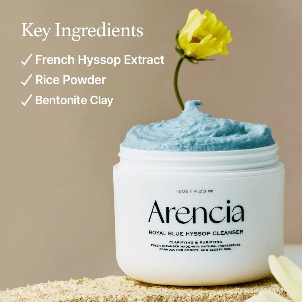 Slika kozarca "Arencia Royal Blue Hyssop Cleanser" napolnjenega z modro čistilno kremo. Rumena roža štrli iz kreme. Besedilo na sliki poudarja tri ključne sestavine: French Hyssop Extract, Rice Powder in Bentonite Clay.