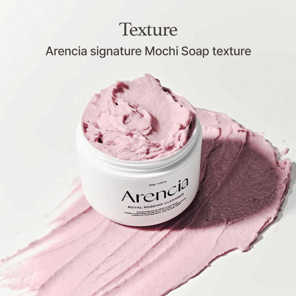 Lonček z Arencia Royal Rosehip Cleanser je viden na razmazanem roza čistilu z mochi podobno teksturo. Besedilo zgoraj opisuje izdelek kot Arenciino podpisno teksturo.