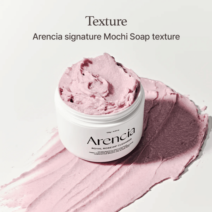 Lonček z Arencia Royal Rosehip Cleanser je viden na razmazanem roza čistilu z mochi podobno teksturo. Besedilo zgoraj opisuje izdelek kot Arenciino podpisno teksturo.