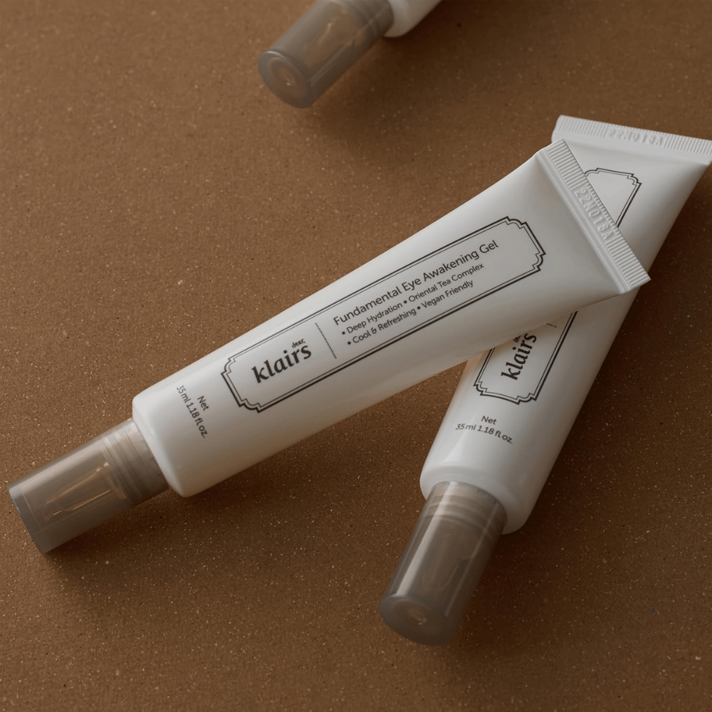 Lifestyle slika z dvema tubama Klairs Fundamental Eye Awakening Gel, postavljenima na toplo rjavo ozadje. Kompozicija poudarja elegantno oblikovanje izdelka in premium občutek, hkrati pa je fokus na negi oči in vlaženju.