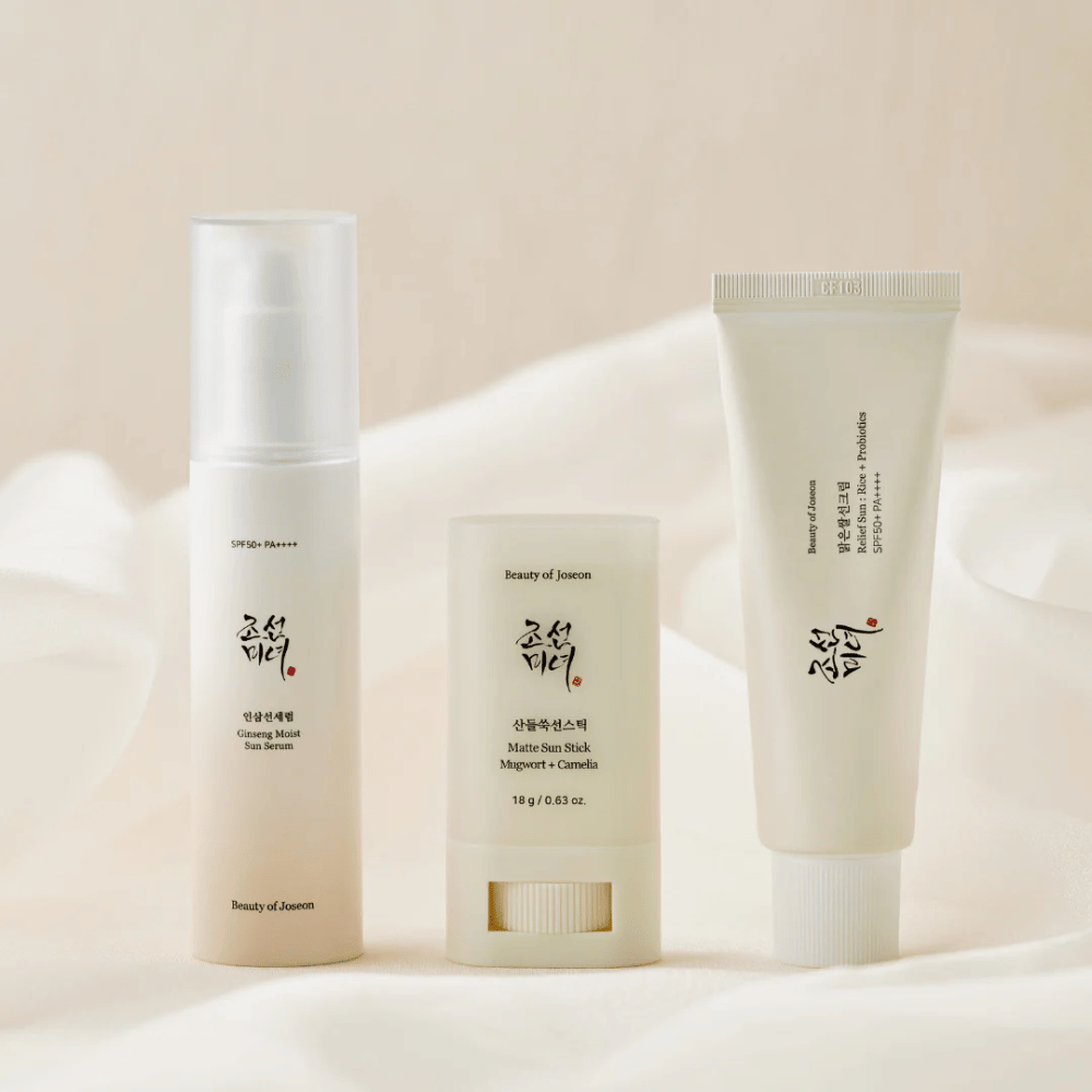 Trije izdelki za zaščito pred soncem iz Beauty of Joseon - Ginseng Moist Sun Serum, Matte Sun Stick in Relief Sun Rice + Probiotics - so postavljeni na kremasto tkanino z mehko svetlobo.