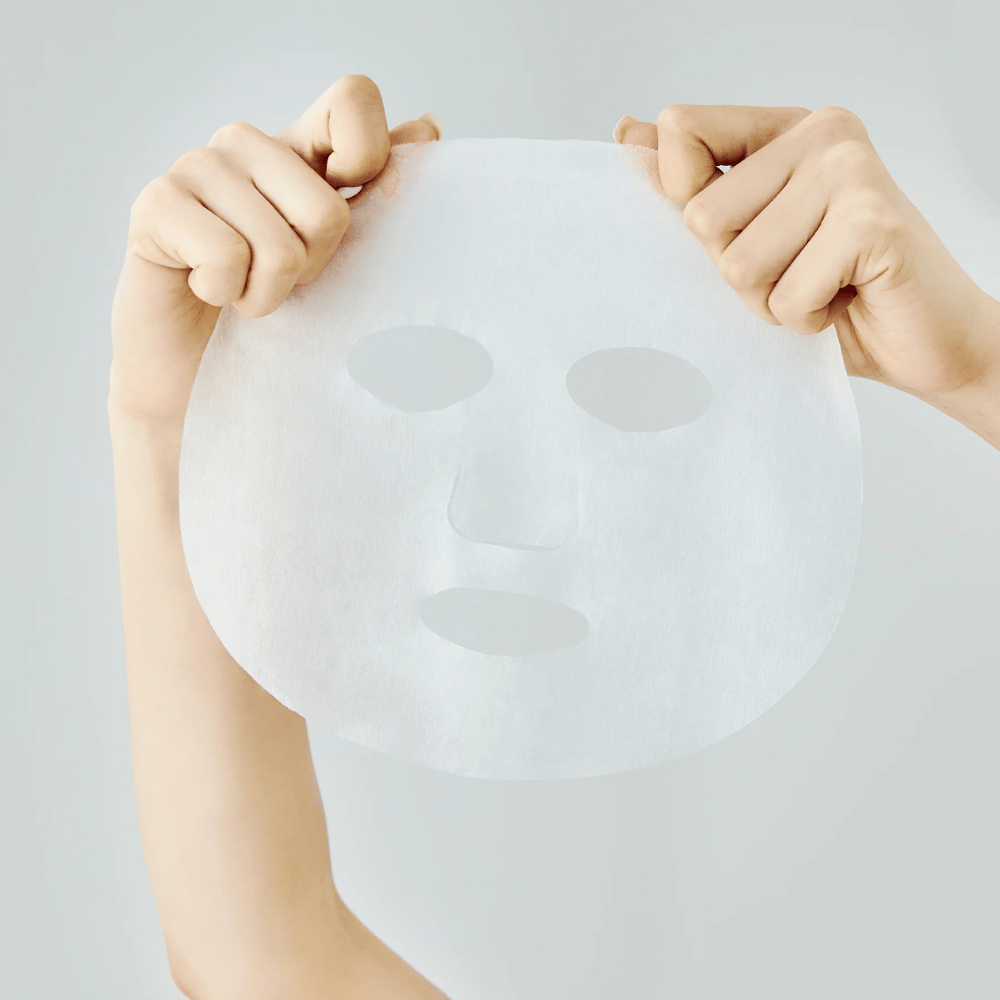 Dve roki držita belo masko proti svetlo sivi podlagi z vidnimi izrezi za oči, nos in usta.