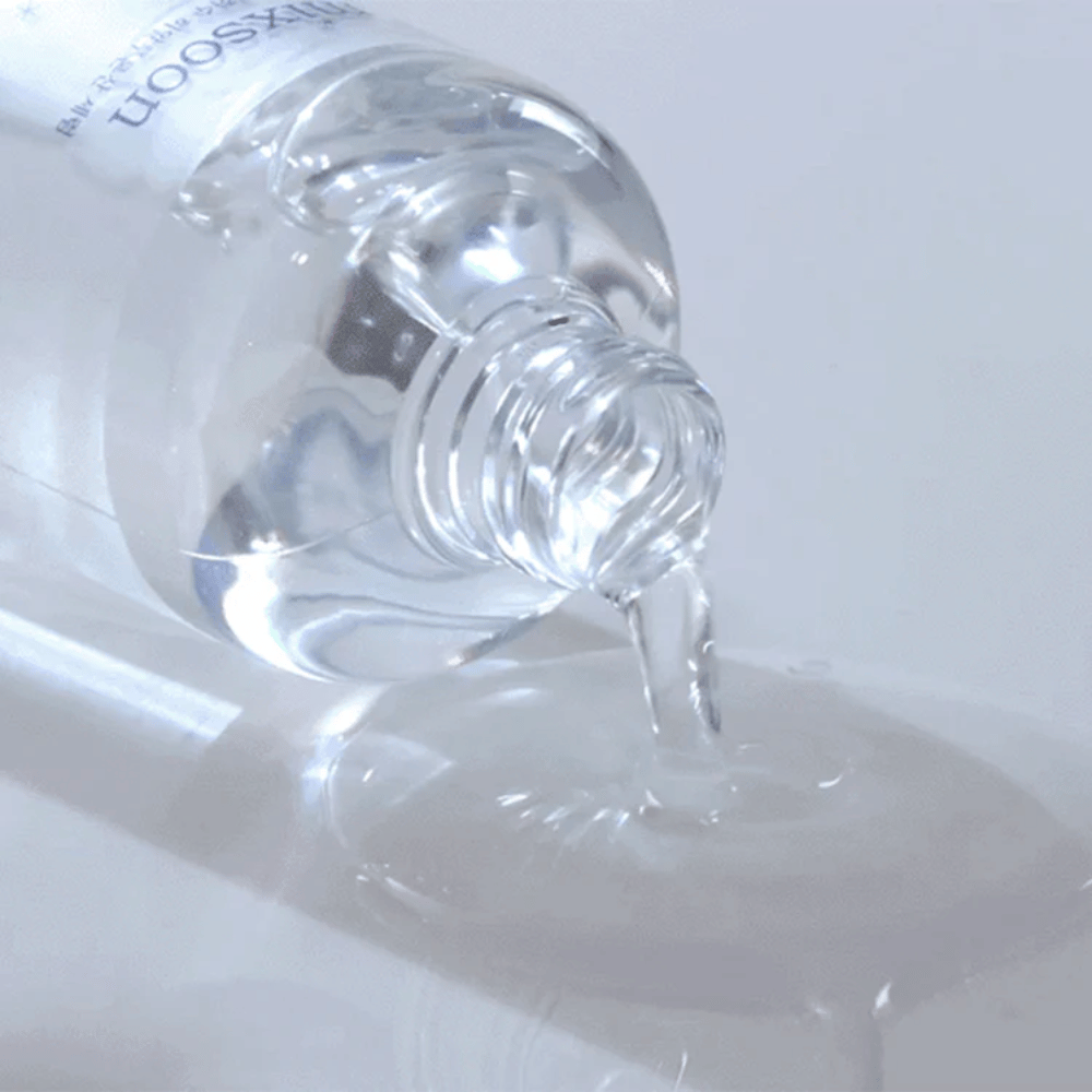 Povečava prozornega seruma, ki se vliva iz stekleničke Mixsoon Glacier Water Hyaluronic Acid Serum na sijočo površino.