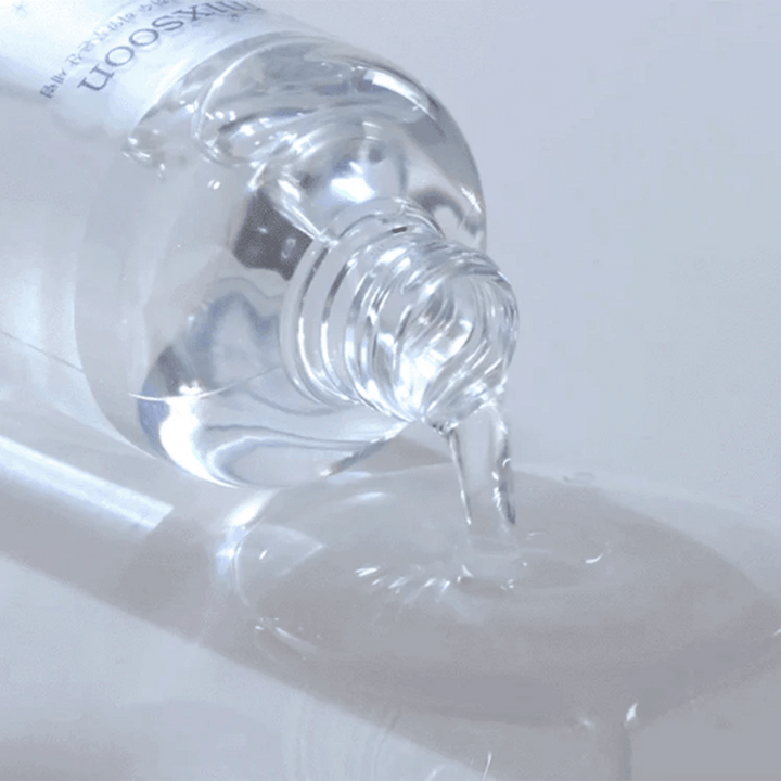 Povečava prozornega seruma, ki se vliva iz stekleničke Mixsoon Glacier Water Hyaluronic Acid Serum na sijočo površino.