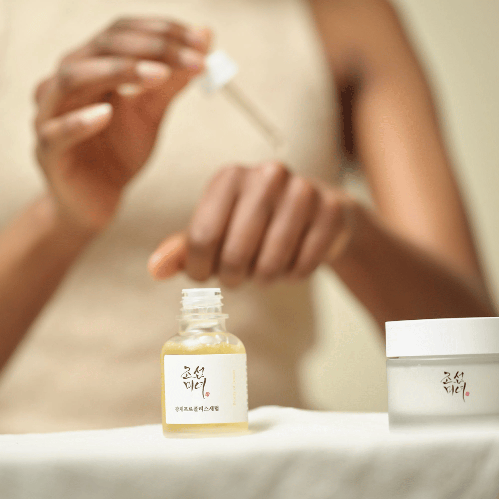 Oseba, ki nanaša Beauty of Joseon Glow Serum s pipeto iz steklene stekleničke, s kozarcem iste serije poleg na mizi.
