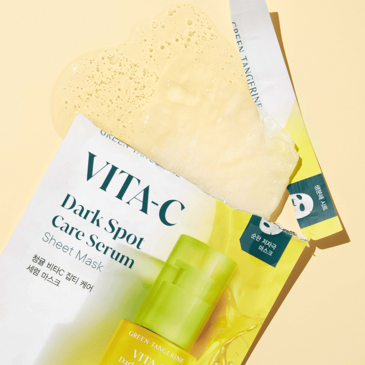 Odprta embalaža VITA-C Dark Spot Care Serum Sheet Mask z obrazno masko na vrhu in zeleno stekleničko seruma, vse na rumenem ozadju.