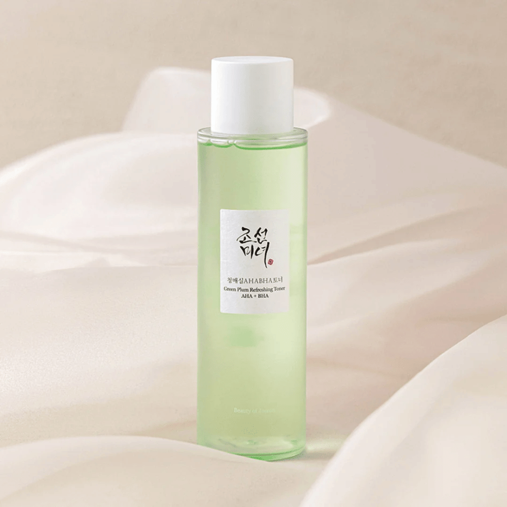Steklenica z "Green Plum Refreshing Toner" AHA + BHA stoji na mehkem, svetlem ozadju. Izdelek ima zeleno tekočino, ki odraža svežino in naravnost. Nalepka je bela s črnimi in rdečimi poudarki, kar daje eleganten in čist videz.