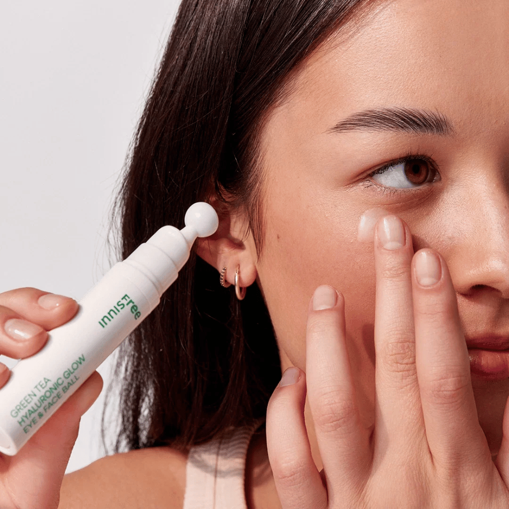 Oseba nanaša Innisfree Green Tea Hyaluronic Glow Eye & Face Ball na kožo pod očmi. Izdelek ima bel, okrogel aplikator in je v beli steklenički z zelenim besedilom. Obraz uporabnika je delno viden in se osredotoča na nanos izdelka.