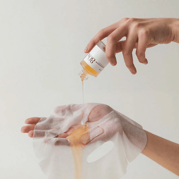 Roka drži zlato, gosto tekočino iz majhne stekleničke z oznako '108' na prozorni obrazni maski, ki jo drži druga roka. Ozadje je nevtralno in enotno, da poudari izdelek.