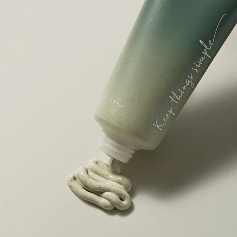 Zelena tuba z napisom "Keep things simple" v beli barvi. Tuba leži na svetli površini in iz nje je iztisnjen zelenkast izdelek z majhnimi črnimi pikami. Videti je kot obrazna maska ali piling izdelek. Na tubi piše 80 ml / 3.52 fl. oz.