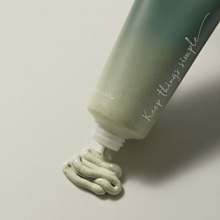 Zelena tuba z napisom "Keep things simple" v beli barvi. Tuba leži na svetli površini in iz nje je iztisnjen zelenkast izdelek z majhnimi črnimi pikami. Videti je kot obrazna maska ali piling izdelek. Na tubi piše 80 ml / 3.52 fl. oz.