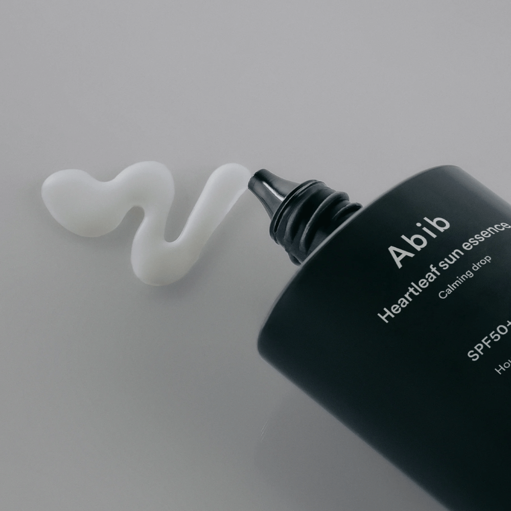 Povečana slika Abib Heartleaf Sun Essence Calming Drop s sončno kremo iztisnjeno v beli, valoviti črti na svetli površini.