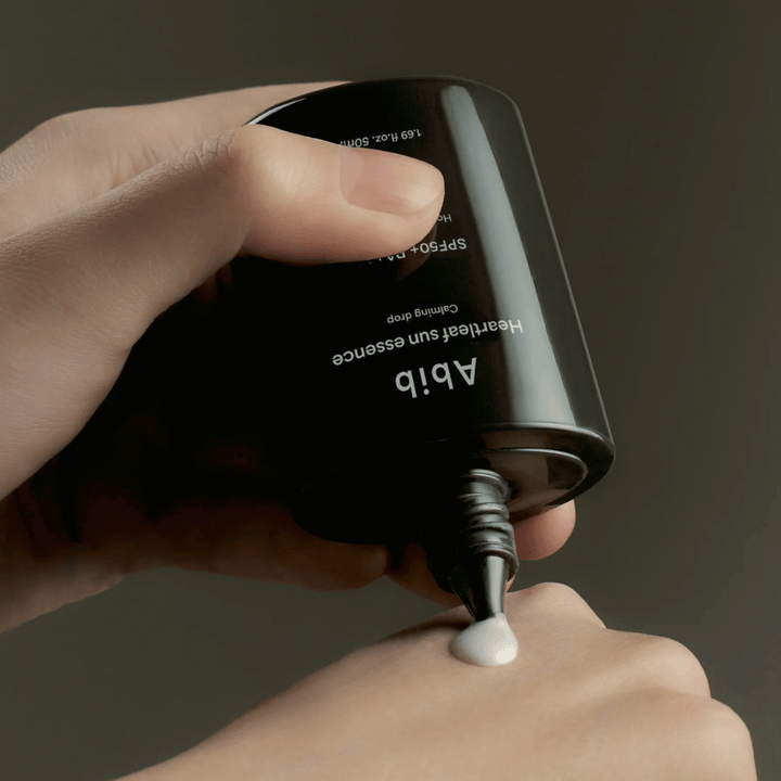 Roka, ki iztisne Abib Heartleaf Sun Essence Calming Drop na zgornji del druge roke, s temnim ozadjem in jasno prikazano teksturo izdelka.