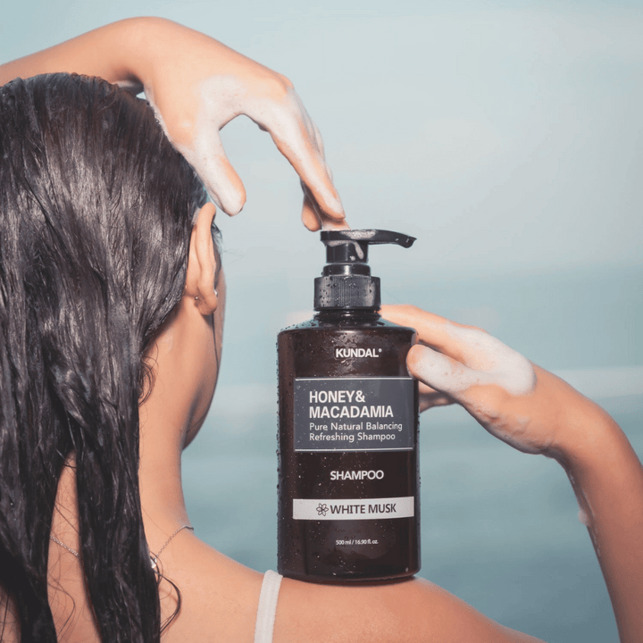 Ženska z mokrimi lasmi nanese peno in drži KUNDAL Honey & Macadamia Nature Shampoo White Musk proti morski kulisi.