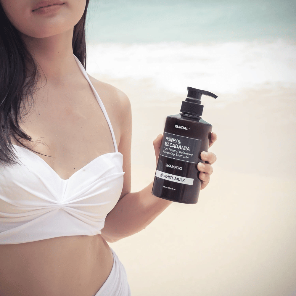 Ženska v belem bikiniju drži KUNDAL Honey & Macadamia Nature Shampoo White Musk na plaži z morjem v ozadju.