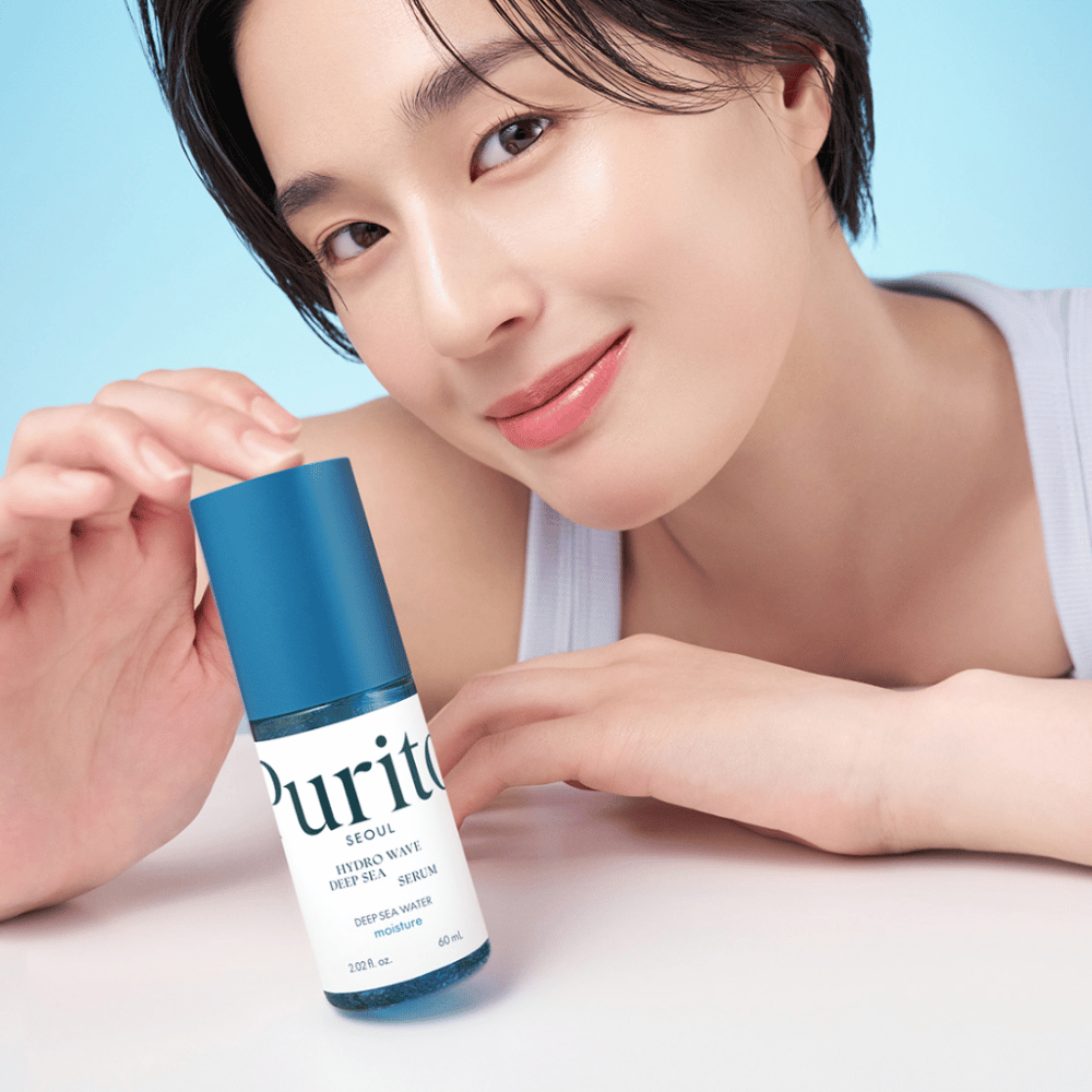 Ženska s kratkimi lasmi drži modri serum Purito, Hydro Wave Deep Sea Serum. Njena koža izgleda sijoča in vlažna, kar poudarja izdelek v ospredju. Ozadje je svetlo modro, kar daje svež in čist občutek. Izdelek je poglobljen v nego kože.
