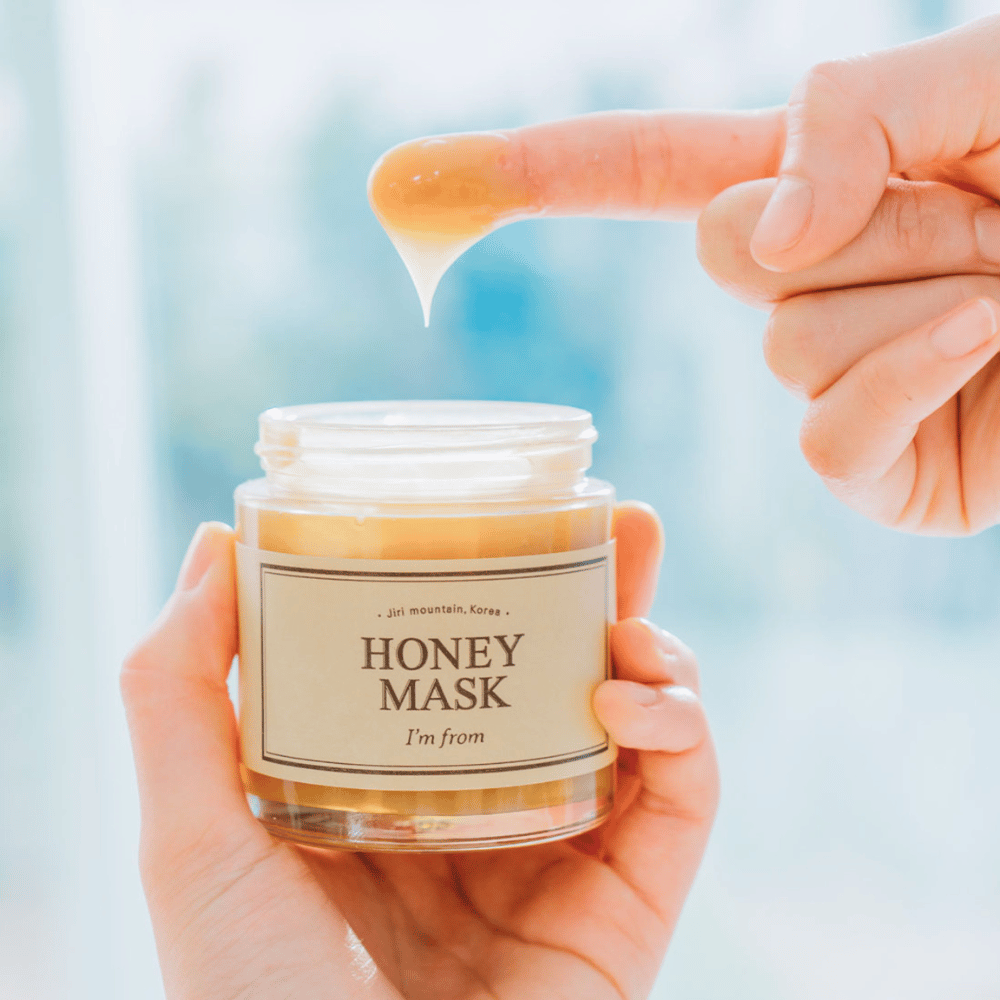 Roka drži odprto posodo "I'm from Honey Mask", in prst se potaplja v zlato masko na svetli podlagi.