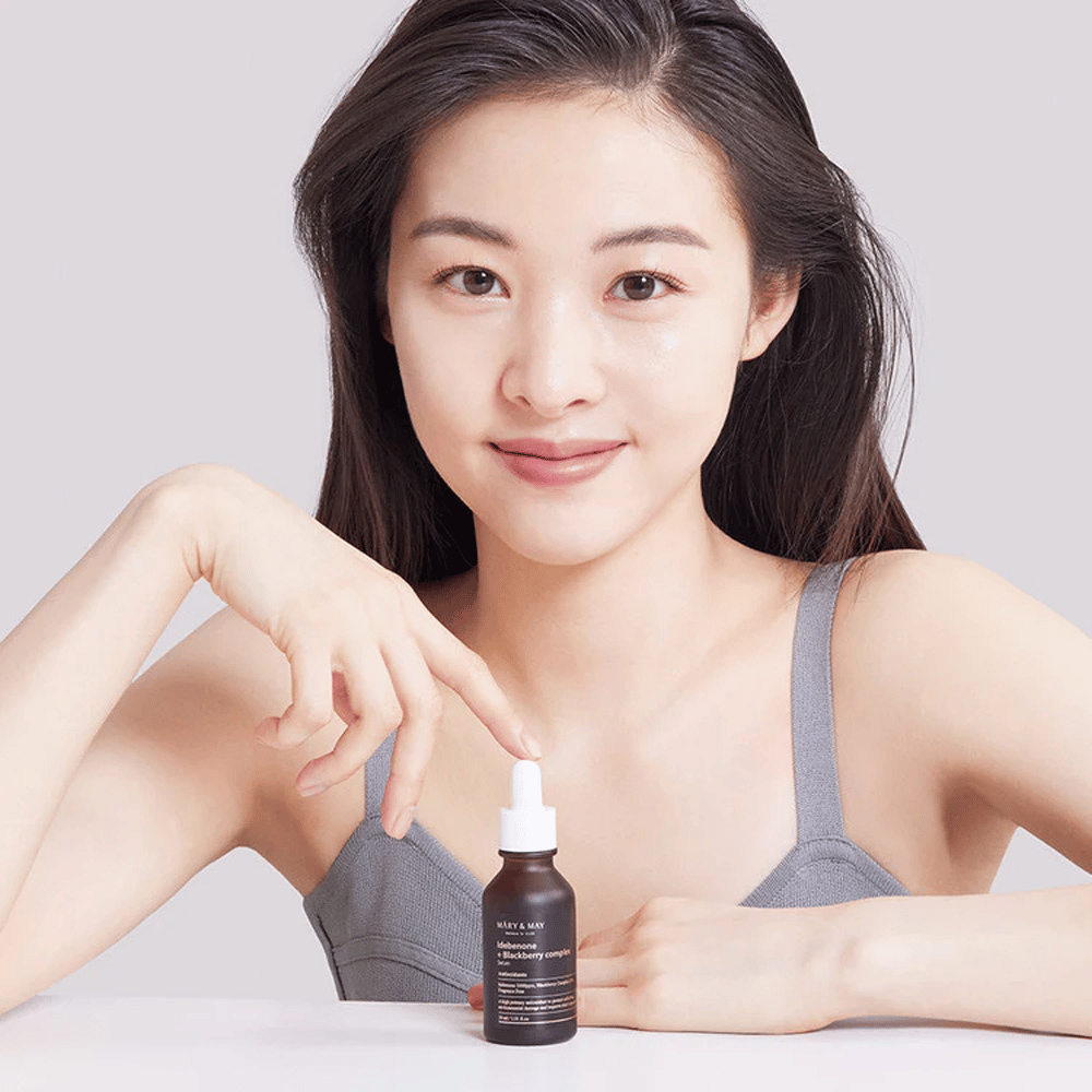 Ženska z dolgimi, temnimi lasmi drži MARY & MAY Idebenone + Blackberry Complex Serum, temno stekleničko s pipeto. Ima vesel nasmeh in s svojo drugo roko kaže na stekleničko. Ženska je oblečena v siv, brezrokavni top in se zdi, da je v svetlem in preprostem okolju, kar poudarja osredotočenost izdelka na nego kože in lepoto.