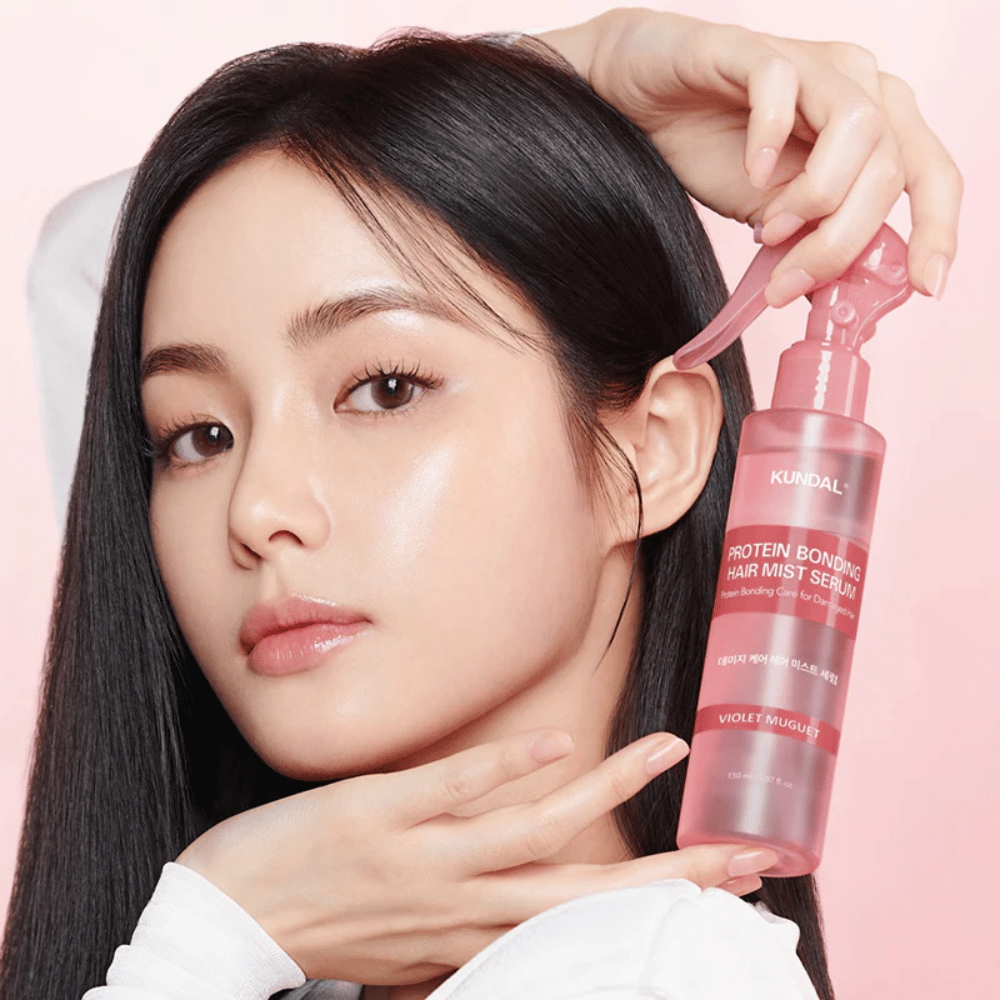 Ženska z dolgimi črnimi lasmi drži KUNDAL Protein Bonding Hair Mist Serum Violet Muguet pred obrazom, na svetlo roza ozadju.