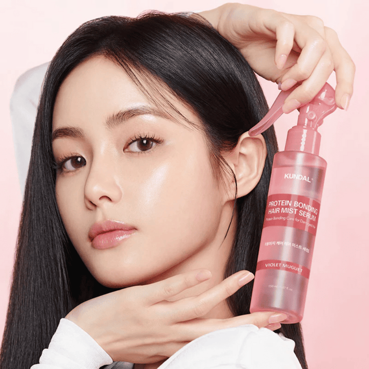 Ženska z dolgimi črnimi lasmi drži KUNDAL Protein Bonding Hair Mist Serum Violet Muguet pred obrazom, na svetlo roza ozadju.