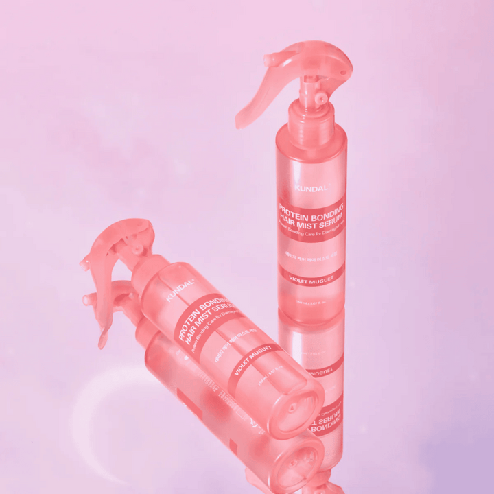 KUNDAL Protein Bonding Hair Mist Serum Violet Muguet v roza razpršilkah, stoječih in ležečih na svetlo roza ozadju z odsevom.