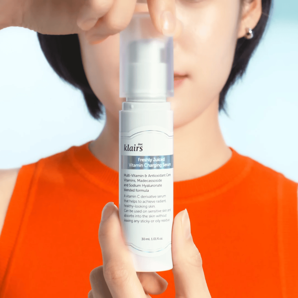 Ženska v oranžni majici drži Klairs Freshly Juiced Vitamin Charging Serum pred seboj na modrem ozadju.