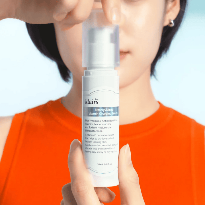Ženska v oranžni majici drži Klairs Freshly Juiced Vitamin Charging Serum pred seboj na modrem ozadju.