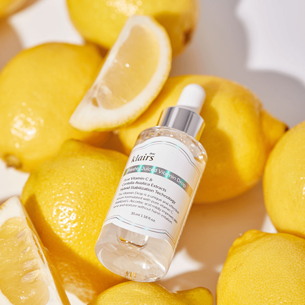 Steklenička seruma Klairs Freshly Juiced Vitamin Drop, obdana s svežimi polovico limone in rezinami limone na svetli površini, poudarja sestavine.
