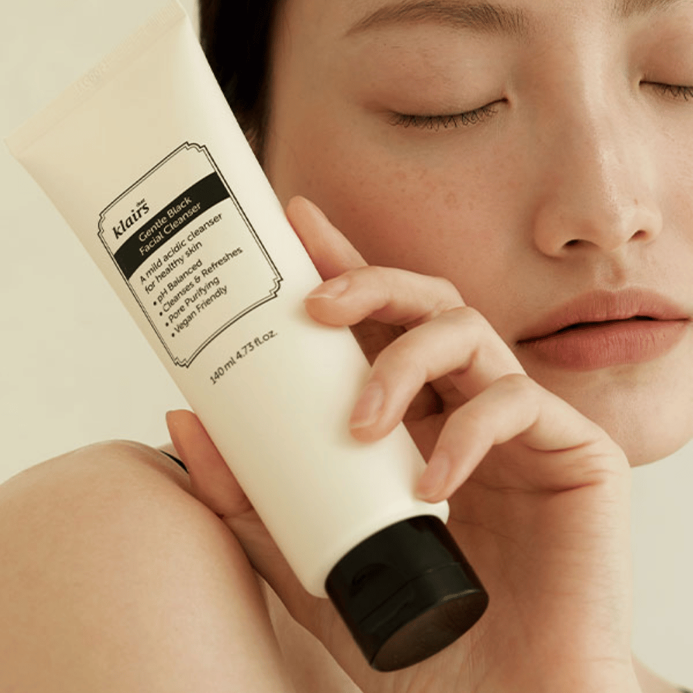 Oseba drži tubo Klairs Gentle Black Facial Cleanser ob svojem licu, zaprte oči in naravni ton kože se vidijo v ozadju.