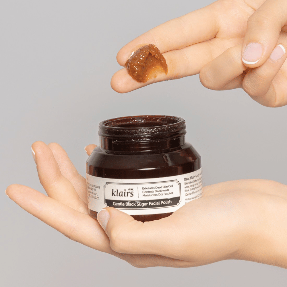 Roke držijo odprt kozarec Klairs Gentle Black Sugar Facial Polish in prikazujejo izdelek na prstu, na sivem ozadju.