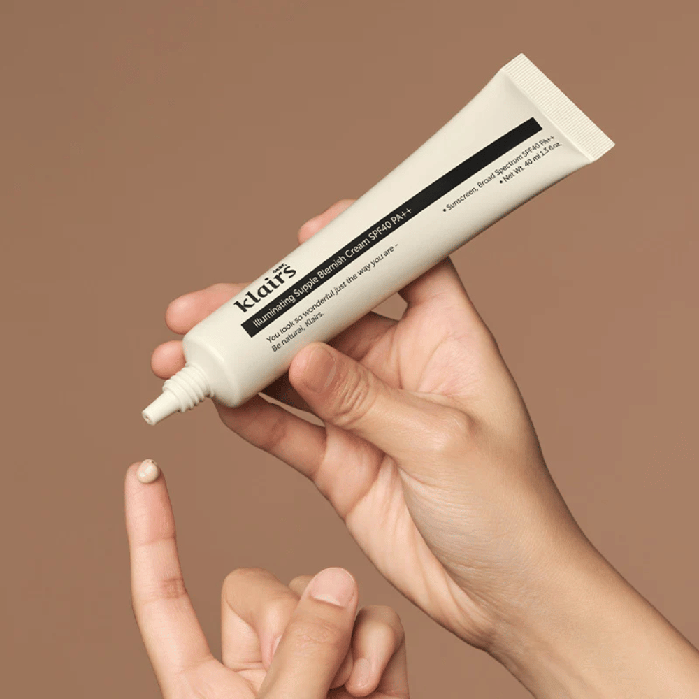 Roka drži tubo Klairs Illuminating Supple Blemish Cream SPF40 PA++ in iztisne majhno količino kreme na prst, na rjavi podlagi.