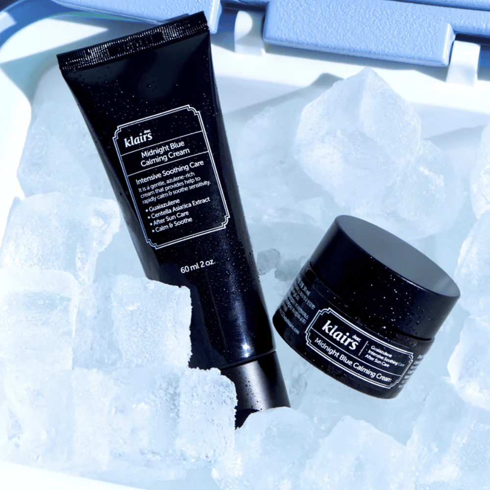 Tuba in posodica Klairs Midnight Blue Calming Cream ležita na ledenih kockah, s poudarkom na pomirjujočih in vlažilnih lastnostih, proti modrem ozadju.