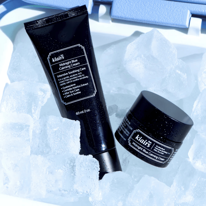 Tuba in posodica Klairs Midnight Blue Calming Cream ležita na ledenih kockah, s poudarkom na pomirjujočih in vlažilnih lastnostih, proti modrem ozadju.