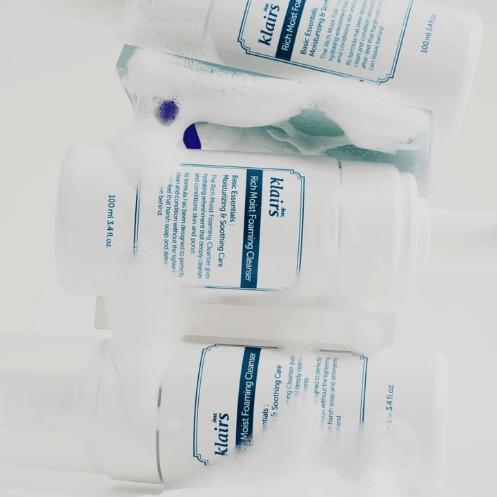 Tri stekleničke Klairs Rich Moist Foaming Cleanser pokrite s peno in kapljicami vode, postavljene na belo površino.