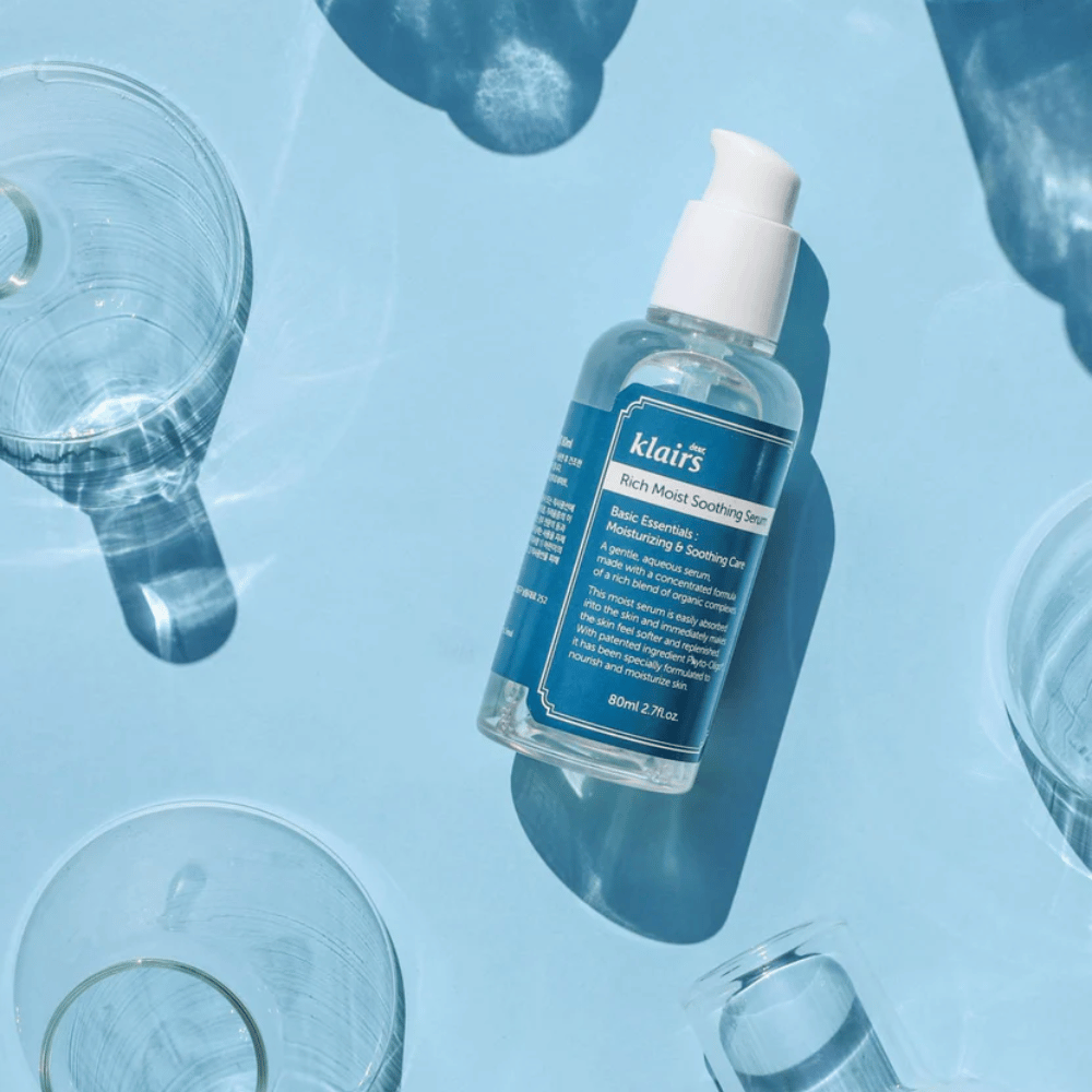 Steklenica Klairs Rich Moist Soothing Serum med kozarci za vodo z odsevi na modri podlagi.