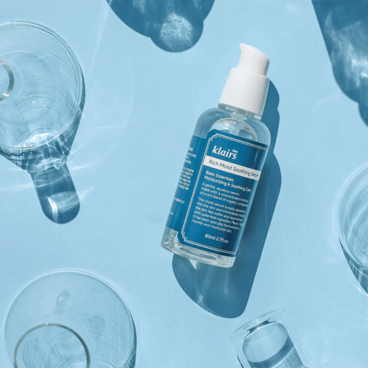 Steklenica Klairs Rich Moist Soothing Serum med kozarci za vodo z odsevi na modri podlagi.