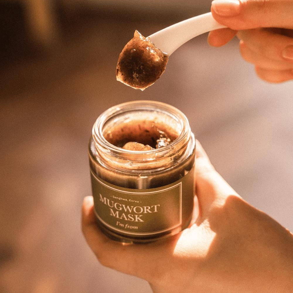 Povečava roke, ki drži I'm From Mugwort Mask, medtem ko žlica dviguje zeliščno gel masko iz kozarca, v topli naravni svetlobi.