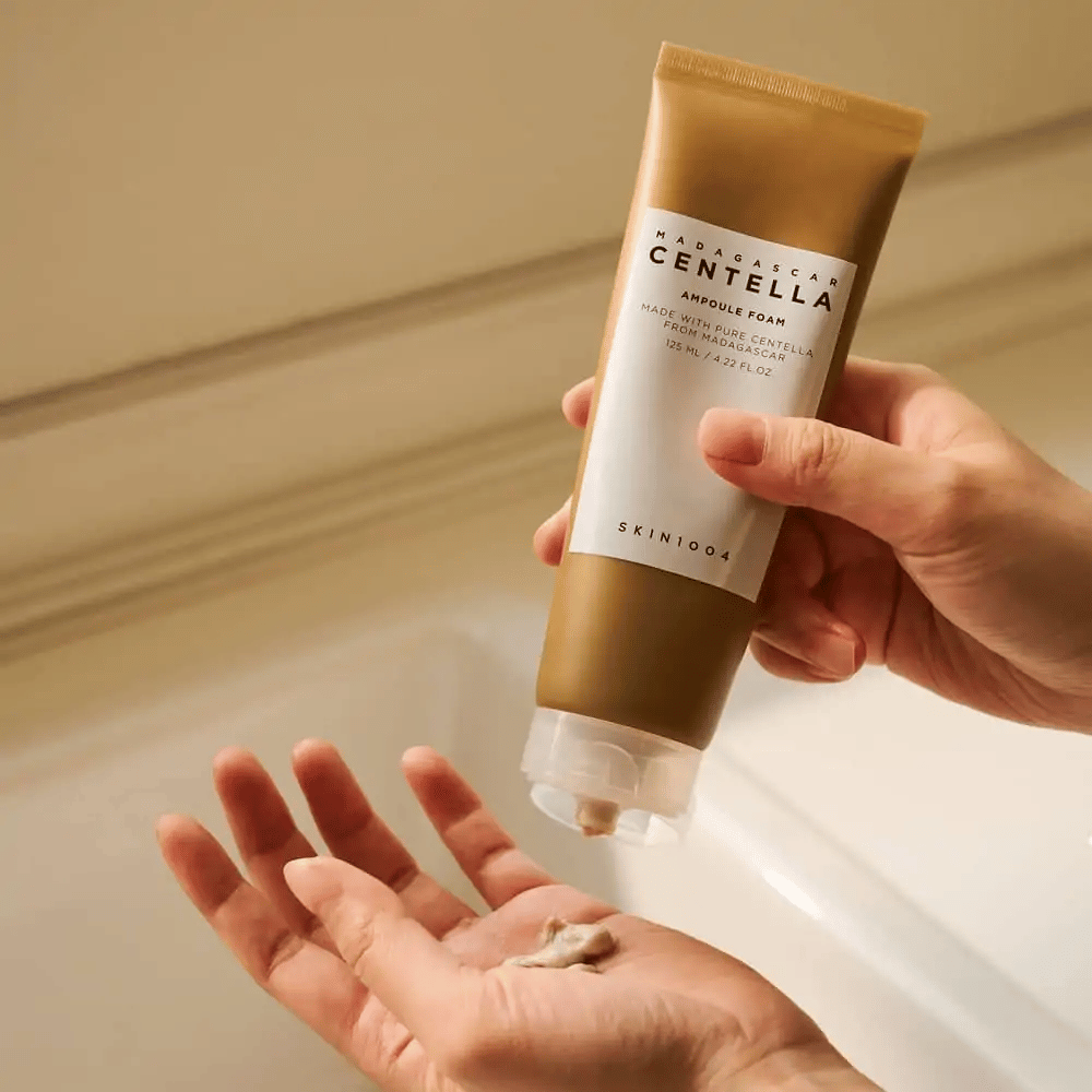 Izdelek na sliki je "Madagascar Centella Ampoule Foam" znamke SKIN1004. To je čistilo za obraz v obliki pene, izdelano iz Centelle z Madagaskarja. Vsebuje 125 ml / 4.22 fl.oz. Roka iztisne majhno količino izdelka za uporabo.