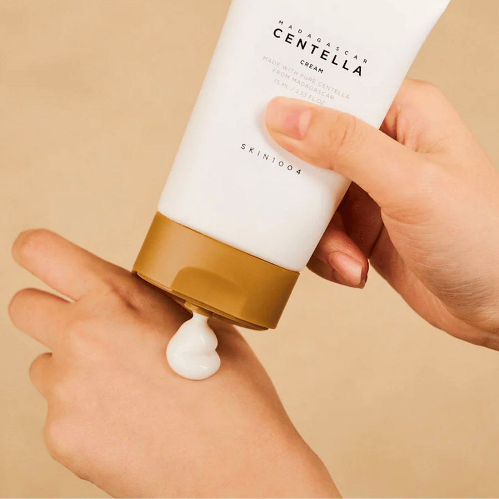 Slika prikazuje osebo, ki drži tubo "Madagascar Centella Cream" od SKIN1004. To je krema za kožo, narejena iz čiste Centelle z Madagaskarja, v velikosti 75 ml / 2,53 fl. oz. Krema se nanaša na roko in je namenjena za hranjenje in vlaženje kože.