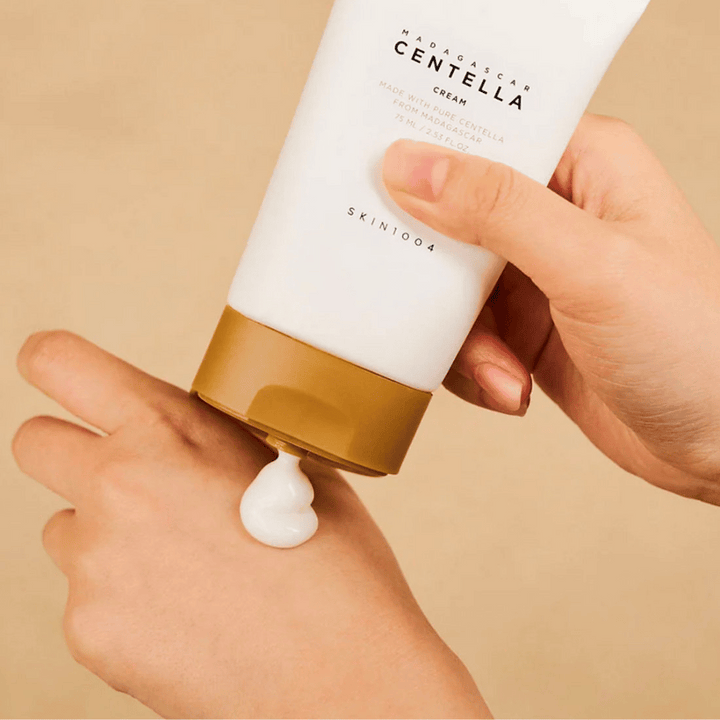 Slika prikazuje osebo, ki drži tubo "Madagascar Centella Cream" od SKIN1004. To je krema za kožo, narejena iz čiste Centelle z Madagaskarja, v velikosti 75 ml / 2,53 fl. oz. Krema se nanaša na roko in je namenjena za hranjenje in vlaženje kože.