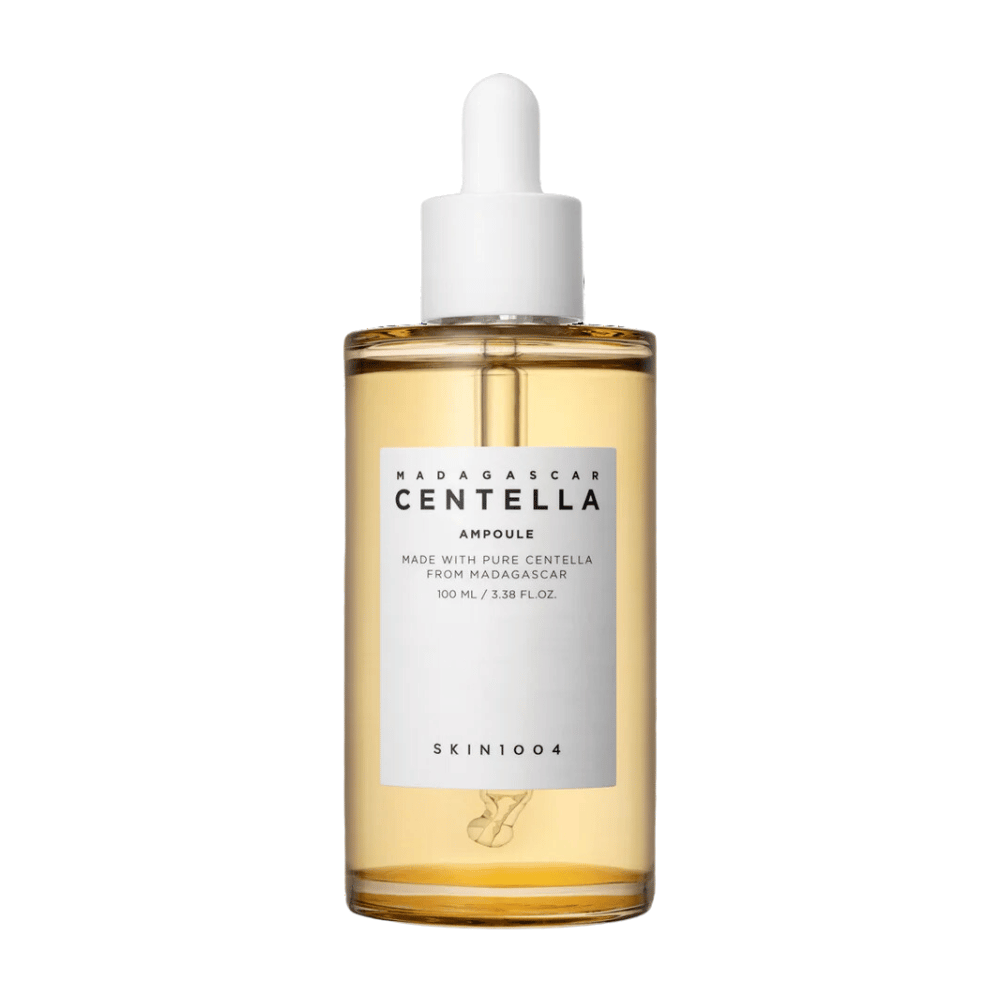 Slika prikazuje steklenico z "Madagascar Centella Asiatica 100 Ampoule" iz SKIN1004. Steklenica vsebuje 100 ml in trdi, da je narejena s čisto centello iz Madagaskarja. Izdelek izgleda kot serum za nego kože, preprosta etiketa pa daje vtis čistosti in naravnosti. Steklenička z kapalko nakazuje, da je to skrbno odmerjen izdelek za nego kože.