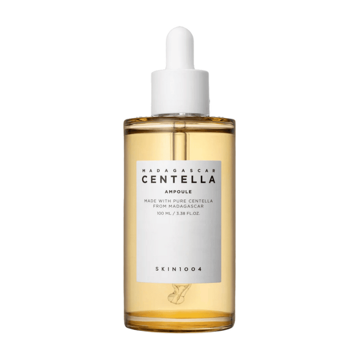 Slika prikazuje steklenico z "Madagascar Centella Asiatica 100 Ampoule" iz SKIN1004. Steklenica vsebuje 100 ml in trdi, da je narejena s čisto centello iz Madagaskarja. Izdelek izgleda kot serum za nego kože, preprosta etiketa pa daje vtis čistosti in naravnosti. Steklenička z kapalko nakazuje, da je to skrbno odmerjen izdelek za nego kože.