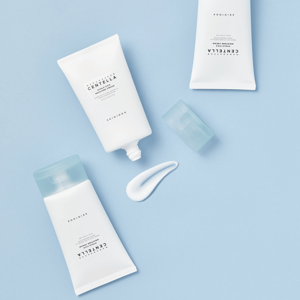 Dve beli tubi s SKIN1004 Centella izdelki za nego kože, kepica izdelka in prozoren moder pokrovček, na svetlo modrem ozadju.