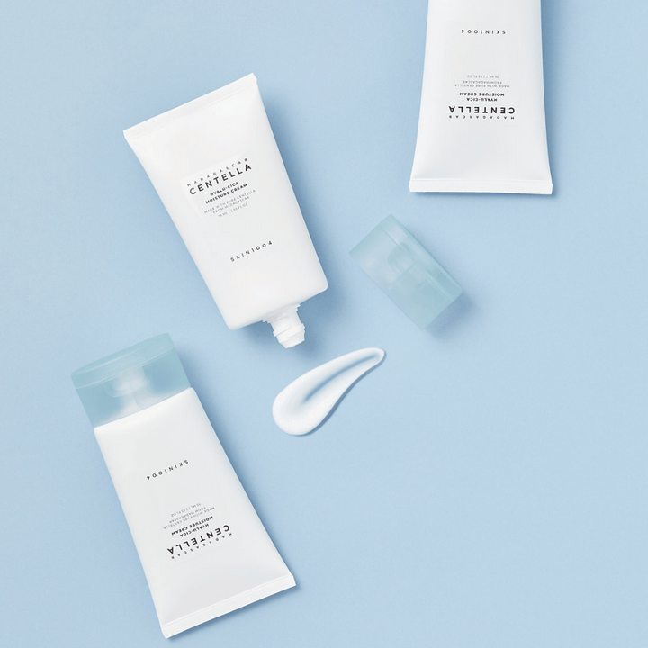 Dve beli tubi s SKIN1004 Centella izdelki za nego kože, kepica izdelka in prozoren moder pokrovček, na svetlo modrem ozadju.