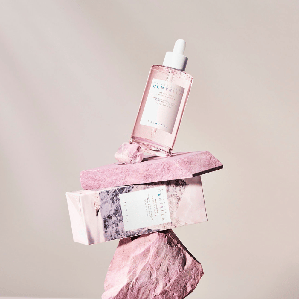 Prozorna steklenička z rožnato ampulo Centella od SKIN1004, uravnotežena na kamnitih blokih, s kartonsko embalažo spodaj, na nevtralni podlagi.